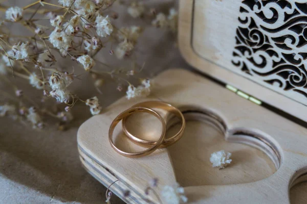 Photo of a wooden box with a pair of gold wedding rings on it. A heart is carved in the box as a symbol of eternal love. Box for rings surrounded by delicate white flowers, the composition is in the background of kraft paper.