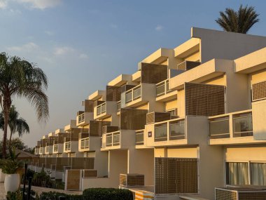 Geometrik balkonları ve doğal ahşap aksanları olan, akşam güneşinin sıcacık parıltısıyla yıkanmış çağdaş beyaz binalar. Palmiye ağaçları sakin tatil beldesi atmosferini çerçeveletip, kıyı zarafetini ve rahatlığını çağrıştırıyor.