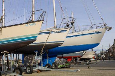 Birkaç yelkenli tekne, açık mavi bir gökyüzünün altında gövdeleri hizalanmış bakım ve kış deposunu gösteren bir liman iskelesinde duruyor..