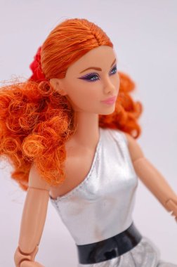 Parlak kırmızı kıvırcık saçlı, modern gümüş bir tulum giyen şık moda bebeği, temiz beyaz bir arka planda, minimal stüdyo görünümlü..