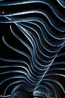 Kıvrımlı mavi metalik ışık dalgaları, karanlık bir arkaplan karşısında, hareket, derinlik ve gelecekteki dijital mimariyi çağrıştıran, ritmik soyut bir yapı oluşturur..