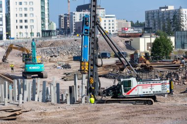 Espoo, Finlandiya - 1 Ağustos 2025: Finnoo 'daki Meritie karayolu şantiyesi, ağır ekipmanlar ve modern altyapı gelişiminin ortasında yere beton yığınları döşeyen güçlü bir kazık makinesi
