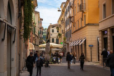 İtalya, Verona - 1 Ekim 2025: Renkli eski binalar, balkonlar ve panjurlarla dolu bir sokaktaki turistler, tarihi şehrin ve zamansız İtalyan atmosferinin cazibesini yakalıyorlar.