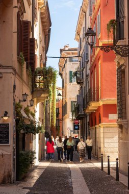 İtalya, Verona - 1 Ekim 2025: Dar bir sokaktaki turistler renkli eski binalar, balkonlar ve panjurlarla dolu, tarihi şehrin ve zamansız İtalyan atmosferinin cazibesini yakalıyorlar.
