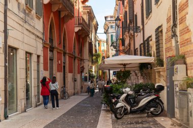 İtalya, Verona - 1 Ekim 2025: Dar bir sokaktaki turistler renkli eski binalar, balkonlar ve panjurlarla dolu, tarihi şehrin ve zamansız İtalyan atmosferinin cazibesini yakalıyorlar.