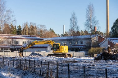 Bir kazıcı, kış koşulları ve sakin, donmuş bir manzarayla çevrili, karla kaplı bir inşaat sahasında boşta duruyor.