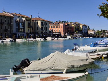 İtalya, Peschiera del Garda - 28 Eylül 2025: Renkli çiçekli yatakları, manzaralı bahçeleri ve kanal boyunca yanaşmış motorlu tekneleri olan manzaralı bir liman.