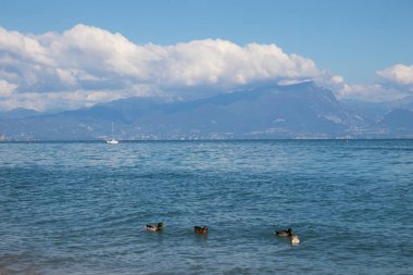 Ördekli Garda Gölü manzarası ve berrak mavi sularda bir yelkenli ve arka planda dağlar, barışçıl doğa ve seyahat atmosferi sergiliyor.