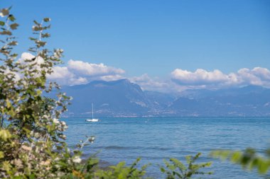 İtalya 'nın en büyük gölü olan Garda Gölü manzaralı arka planda barışçıl doğa ve seyahat atmosferini gösteren berrak mavi sularda ve dağlarda bir yelkenli.