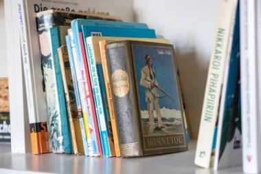 Almanca ve Fince birkaç ciltli ve karton kaplı raf. Samimi, eğitici iç mekan okuma, edebiyat ve çok dilli koleksiyonları vurguluyor
