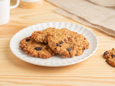 Taze pişmiş yulaf ezmeli kuru üzümlü kurabiyeler ahşap bir masada beyaz flüt tabağında. Çiğnenmiş, yulaf, kuru üzüm ve klasik pişirme malzemeleriyle yapılan tatlı ikramlardan.