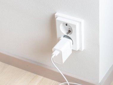 Çift elektrik prizinin yakın çekimi beyaz bir duvara monte edilmiş, prizlerden birine şarj cihazı takılmış. Elektrik tüketimi ve enerji tasarruflarının farkında olmak, elektrik kullanımının yönetilmesi