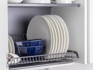 Parlak metal bir bulaşık kurutma askısı modern bir mutfaktaki duvar dolabına düzgünce yerleştirilir. Askıda beyaz seramik mutfak gereçleri, tabaklar ve kupalar var. Hepsi düzenli olarak düzenlenmiş.