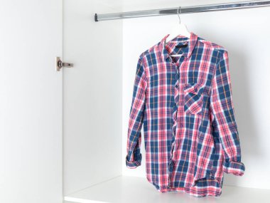 Kırmızı, beyaz ve mavi gömlekli, beyaz bir gardıroptaki ahşap bir askıya sarılı. Günlük pamuk gömlek.