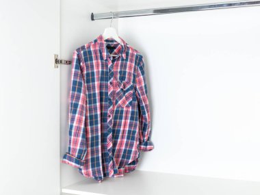 Kırmızı, beyaz ve mavi gömlekli, beyaz bir gardıroptaki ahşap bir askıya sarılı. Günlük pamuk gömlek.