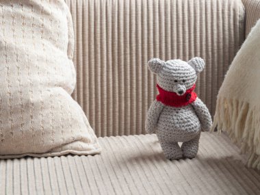 Bej renkli kadife koltukta, krem rengi yastık ve beyaz battaniyenin yanında duran kırmızı eşarplı el yapımı gri bir oyuncak ayı. Bebek odası ya da hediye olarak samimi amigurumi oyuncağı.