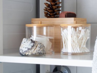 Pamuk ve bezle kaplı bambu kapaklı kavanozlar, dekoratif deniz kabuğu ve kozalak, terakotta sabunu ve tüylü beyaz havlu içeren beyaz banyo rafı. Düzenli ve temiz.