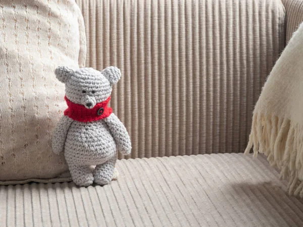 Bej renkli kadife koltukta, krem rengi yastık ve beyaz battaniyenin yanında duran kırmızı eşarplı el yapımı gri bir oyuncak ayı. Bebek odası ya da hediye olarak samimi amigurumi oyuncağı.