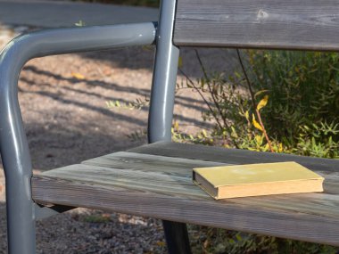 Modern ahşap ve metal bir bankta duran küçük soluk sarı bir kitap. Çakıl taşları, sonbahar yeşillikleri ve gölgeler ile güneşli park manzarası, sessiz okuma anını çağrıştırıyor.