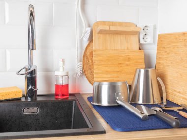 Karanlık bir lavabonun etrafında iyi organize edilmiş bir mutfak tezgahında krom çekiş musluğu, temizlik malzemeleri, bambu kesme tahtaları ve fırça, paslanmaz çelik kaplar ve beyaz bir döşeme serpintisi bulunur.