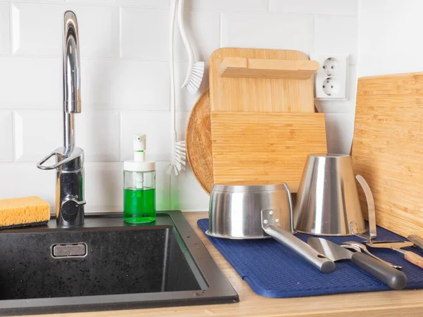 Karanlık bir lavabonun etrafında iyi organize edilmiş bir mutfak tezgahında krom çekiş musluğu, temizlik malzemeleri, bambu kesme tahtaları ve fırça, paslanmaz çelik kaplar ve beyaz bir döşeme serpintisi bulunur.