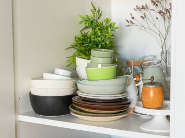 Mutfak rafında çeşitli seramik tabaklar, kaseler ve yeşil saksı bitkisi ve ahşap baharat değirmeninin yanında nötr renklerde bardaklar var.