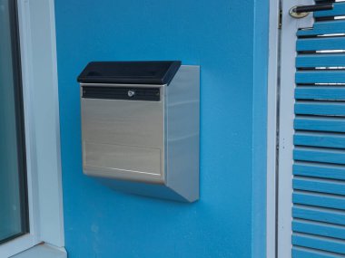 Siyah kapaklı gümüş metal bir posta kutusu beyaz hoparlör kapısının yanındaki parlak mavi bir duvara monte edilmiştir.