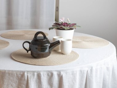 Seramik bir çaydanlık, iki beyaz fincan ve bir saksı Afrika menekşesi ile çevrili, beyaz kaplı bir masanın üzerinde yuvarlak bir plasemat üzerinde oturur. Sahne sıcak bir çay saatine benziyor.