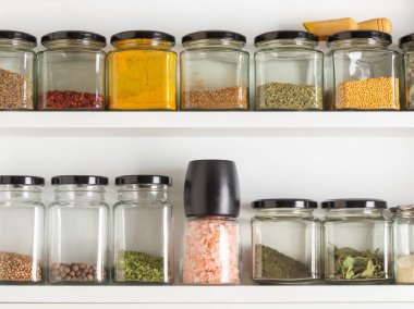 İki beyaz raf çeşitli baharat ve otlarla dolu düzenli bir cam kavanoz koleksiyonu sergiliyor. Bu da düzenli bir mutfak, yemek pişirme tutkusu ve pratik bir yerli organizasyon olduğunu gösteriyor.