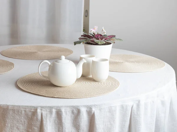 Seramik bir çaydanlık, iki beyaz fincan ve bir saksı Afrika menekşesi ile çevrili, beyaz kaplı bir masanın üzerinde yuvarlak bir plasemat üzerinde oturur. Sahne sıcak bir çay saatine benziyor.