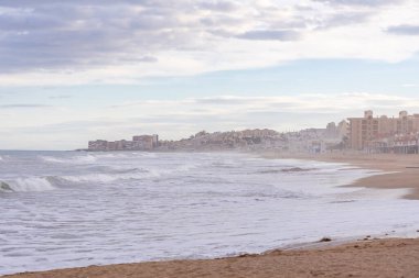 İspanya, Torrevieja 'da sakin bir sahil manzarası, yumuşak, bulutlu bir gökyüzü altında kumlu kıyıya vuran hafif dalgalar ve Akdeniz boyunca uzanan bir kıyı kasabası..