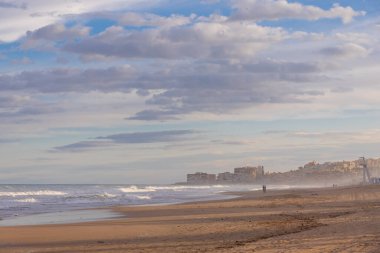 İspanya, Torrevieja 'da sakin bir sahil manzarası. Hafif dalgalar bulutlu bir gökyüzünün altında kuma vuruyor. Uzakta görünen bir kıyı kasabası var..