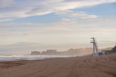 İspanya, Torrevieja 'da sakin bir sahil sahnesi, kumların üzerinde bir cankurtaran kulesi, dalgaların nazikçe çarptığı ve yumuşak bir gökyüzünün altında uzak bir şehir manzarası.