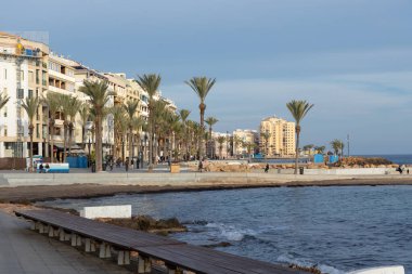 Torrevieja, İspanya - 12 Ocak 2026: Güneşli bir gün Torrevieja 'nın pitoresk kıyı şeridini aydınlatıyor. Derin mavi Akdeniz, kayalık bir dalgakırana ve kumlu bir plaja doğru yavaşça döner.