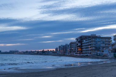 Torrevieja, İspanya - 13 Ocak 2026: Sakin Akdeniz kıyı şeridi alacakaranlığın yumuşak, solan ışığında yıkandı. Yumuşak dalgalar kumlu sahili öperek derin mavi denize karşı köpüklü beyaz bir çıkıntı yaratır. Yukarıdaki gökyüzü gelişen bir tuval.