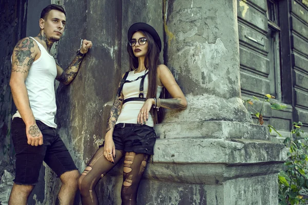 Jovem elegante casal tatuado em shorts pretos em pé na coluna da velha casa arruinada — Fotografia de Stock