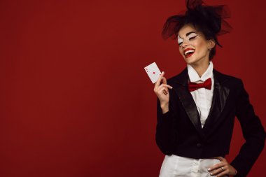 Portrait of a young beautiful lady croupier in an image of joker holding an ace card and laughing clipart