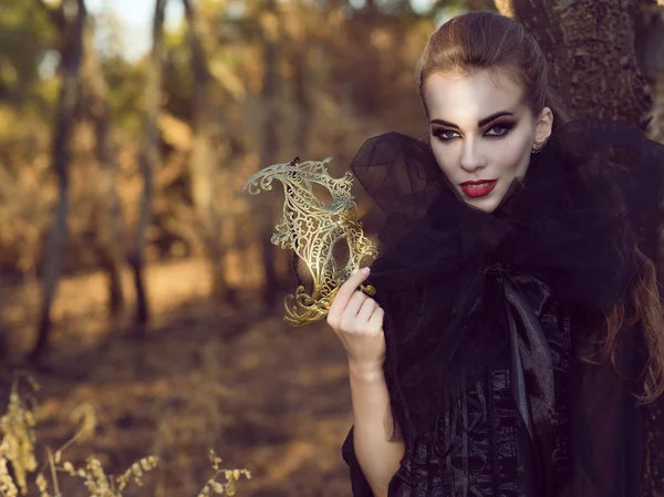 Retrato de mujer vampiro peligroso elegante en el bosque sosteniendo delicada máscara y mirando directamente con mirada depredadora . — Foto de Stock