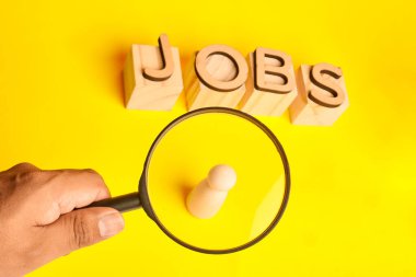 Yakınındaki iş engelleri ile tek bir ahşap figüre odaklanan büyüteç, rekabetçi bir istihdam ortamındaki hedef alımı ve kusursuz yetenek arayışını sembolize ediyor.