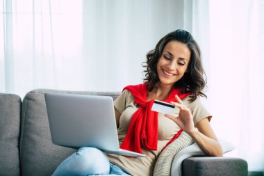 Günlük giysiler içinde güzel mutlu genç bir kadın evdeki kanepede oturuyor ve Kara Cuma 'da online alışveriş yapıyor..