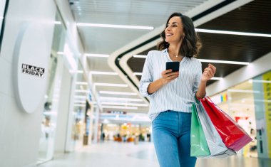 Parlak renkli alışveriş çantalarıyla şık giyinmiş modern, muhteşem, genç bir bayan alışveriş merkezinde yürürken akıllı telefonunu kullanıyor.
