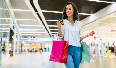 Parlak renkli alışveriş çantalarıyla şık giyinmiş modern, muhteşem, genç bir bayan alışveriş merkezinde yürürken akıllı telefonunu kullanıyor.