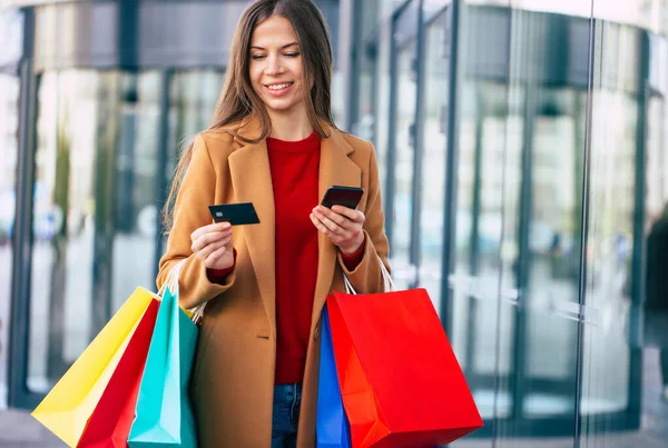 현대의 아름다운 여성이 온라인 뱅킹과 쇼핑백에 스마트폰을 사용하며 쇼핑몰을 야외를 — 스톡 사진