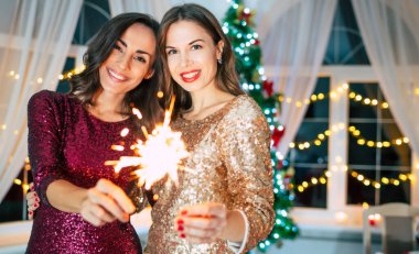 Akşam elbiseli iki güzel kız arkadaş Noel ağacı arka planında poz veriyorlar ve yeni yıl partisinde eğleniyorlar.