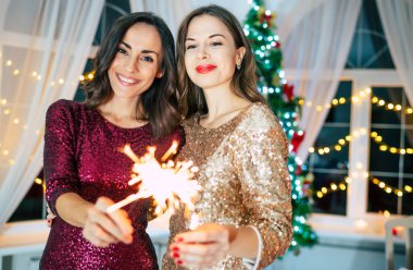 Akşam elbiseli iki güzel kız arkadaş Noel ağacı arka planında poz veriyorlar ve yeni yıl partisinde eğleniyorlar.