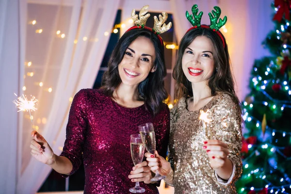Akşam elbiseli iki güzel kız arkadaş Noel ağacı arka planında poz veriyorlar ve yeni yıl partisinde eğleniyorlar.
