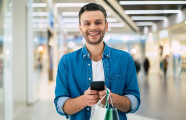 Yakışıklı, genç, şık, sakallı adam siyah cuma günü alışveriş merkezinde yürürken akıllı telefon kullanıyor.