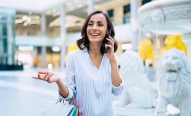 Alışveriş çantalı mutlu, genç, şık bir kadın siyah cuma günü alışveriş merkezinde yürürken akıllı telefon kullanıyor.