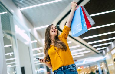 Alışveriş çantalı mutlu, genç, şık bir kadın siyah cuma günü alışveriş merkezinde yürürken akıllı telefon kullanıyor.