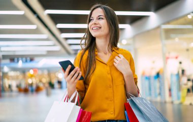 Alışveriş çantalı mutlu, genç, şık bir kadın siyah cuma günü alışveriş merkezinde yürürken akıllı telefon kullanıyor.